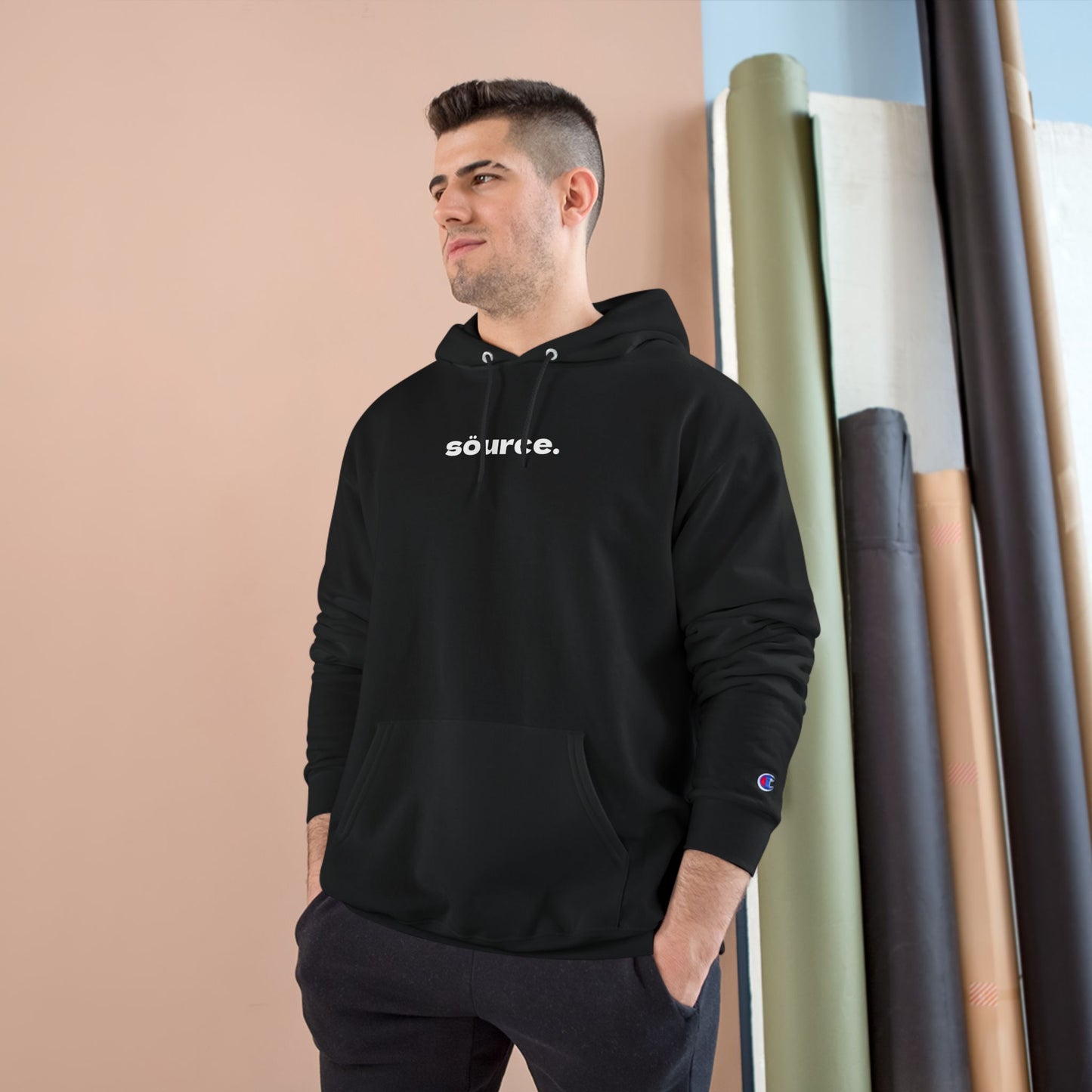 Man wearing a black hoodie with 'söurce.' text, standing against a beige wall with fabric rolls.