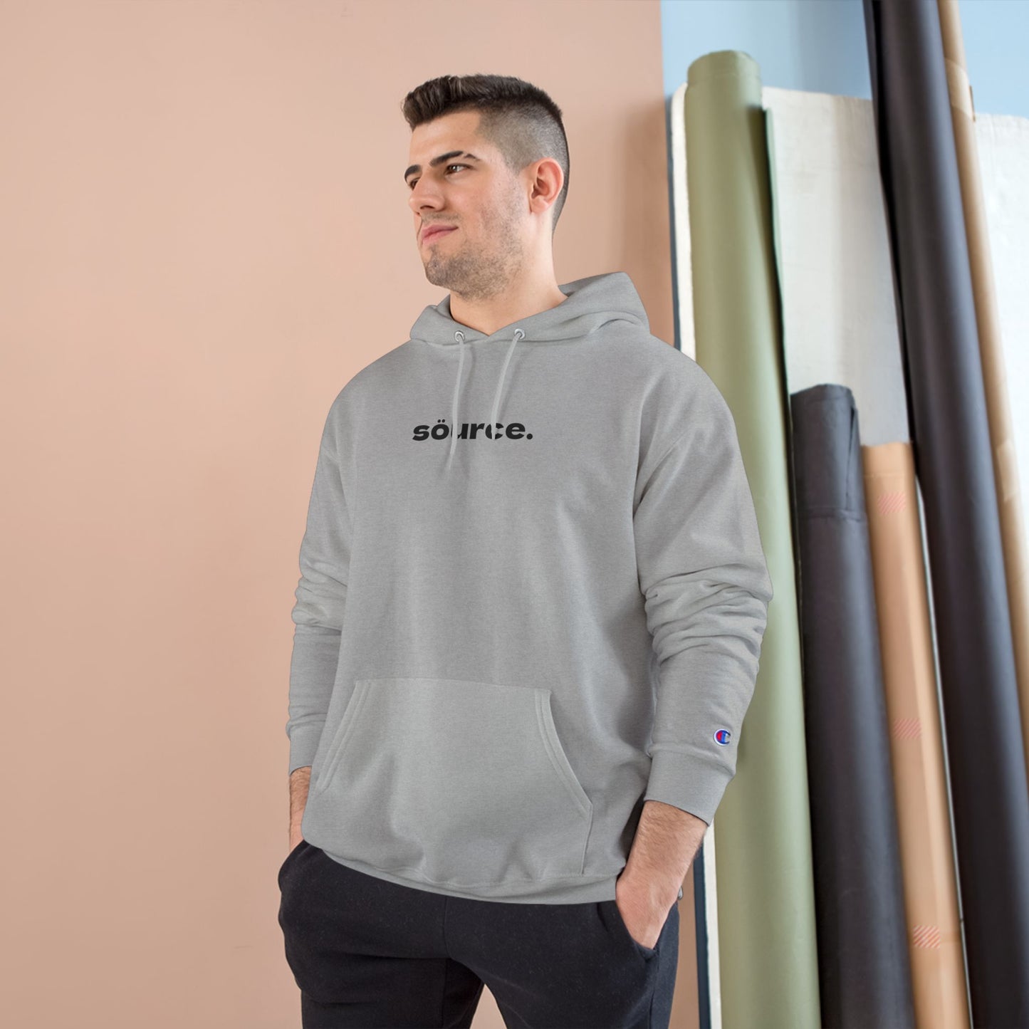 Man wearing a gray hoodie with 'source.' text, standing against a beige wall with rolled-up rugs.