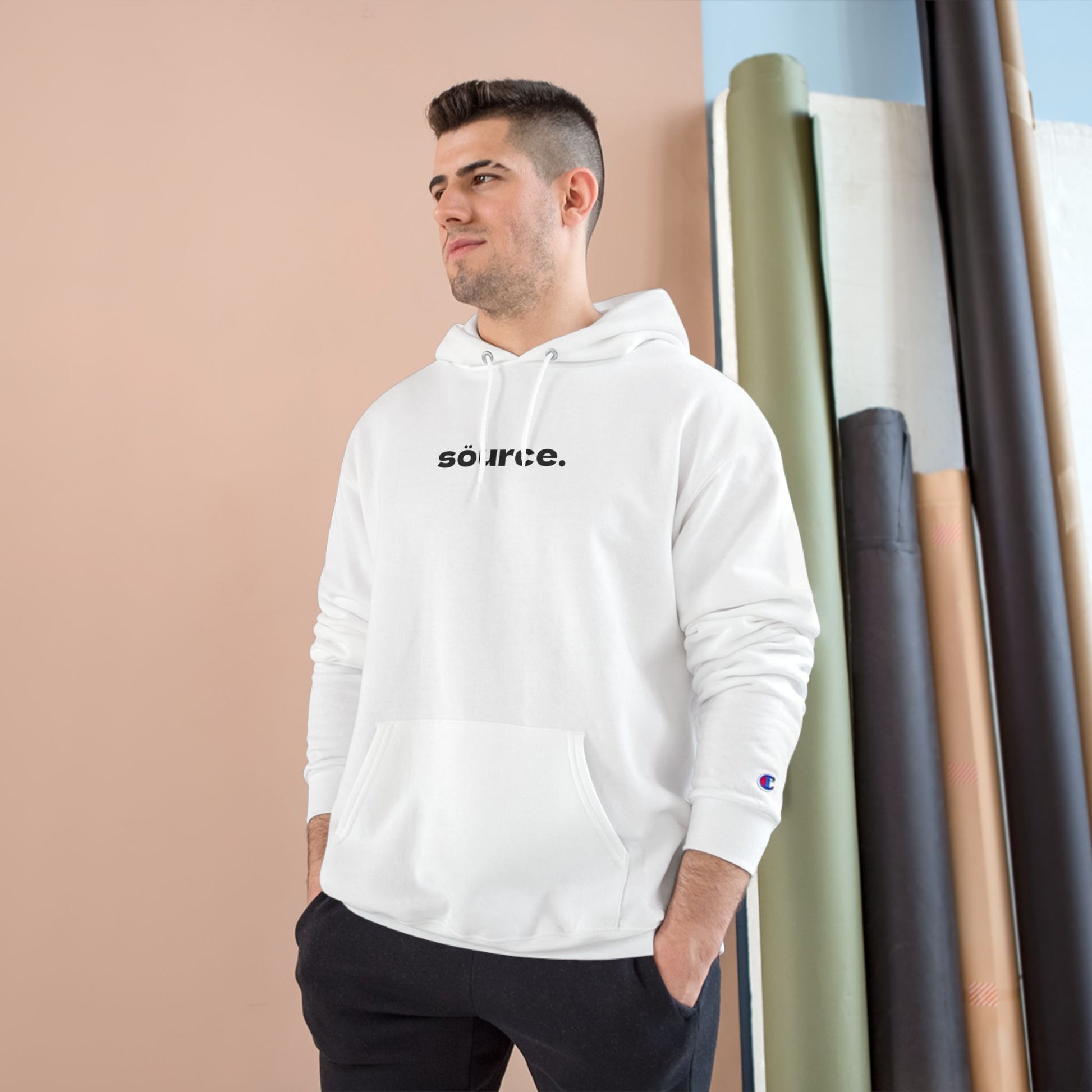 Man wearing a white hoodie with 'source' text, standing against a beige wall with fabric rolls in the background.