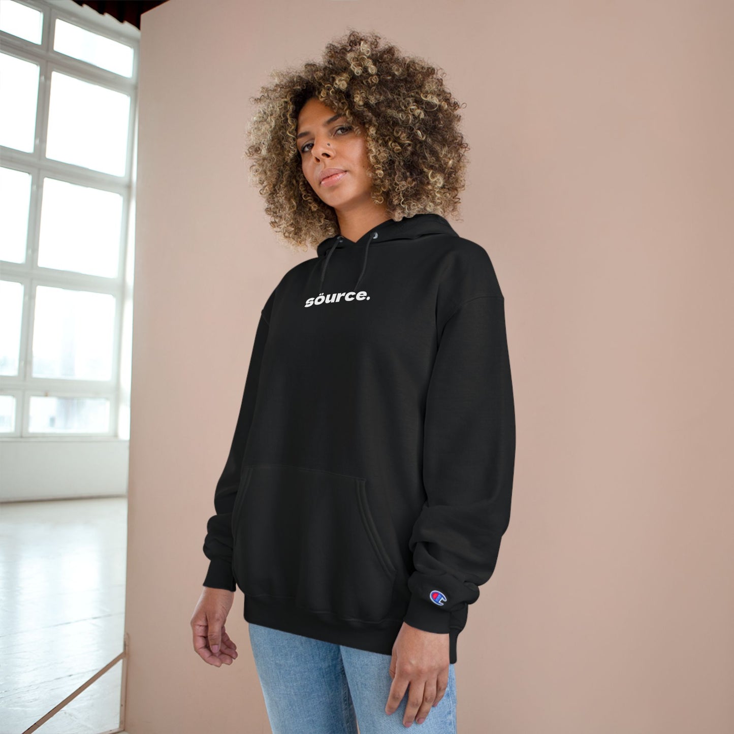 Person wearing a black hoodie with 'source.' text, standing against a beige wall.