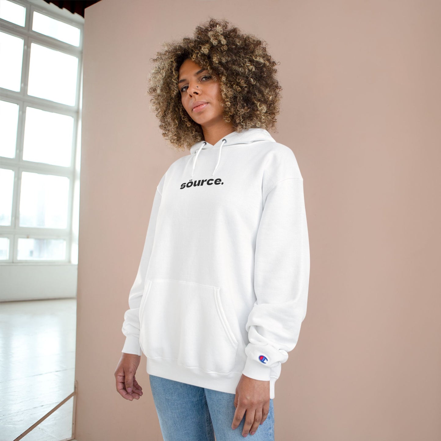 Person wearing a white hoodie with 'source.' text, standing against a beige wall.