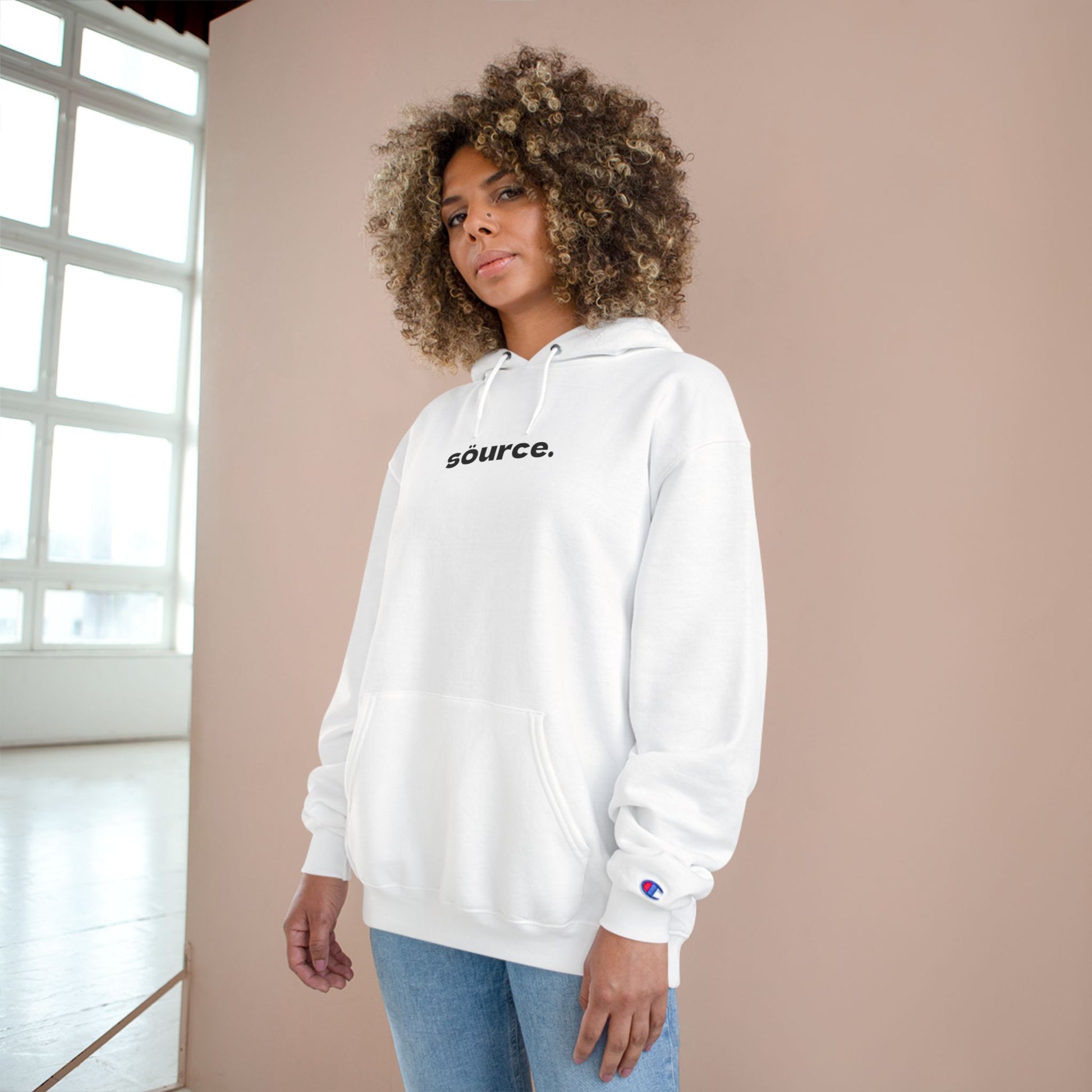 Person wearing a white hoodie with 'source.' text, standing against a beige wall.